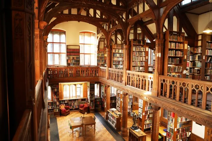 The wooden interior of the library shines in the golden sun seeping through the windows in the background. Many books line the walls of the two story interiors, with a table sitting on the floor below.