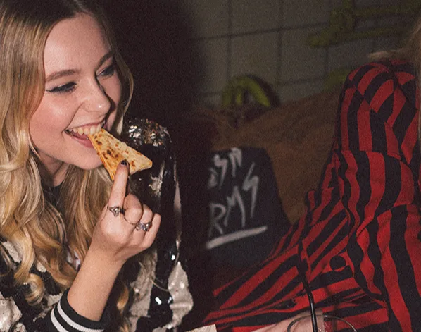 Person with blonde hair in sequined jacket enjoying pizza at a restaurant, with food and drinks on the table.