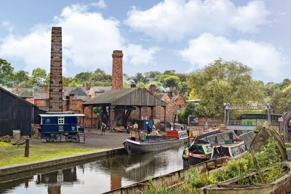 A canal with boats on next to buildings with old-fashioned brick chimneys and an open barn with a wooden roof. 