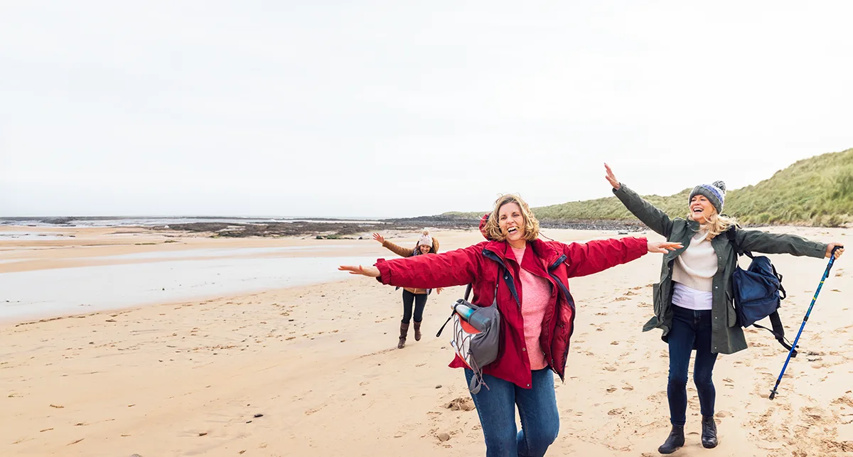 Three white women wearing outdoor gear and waving their arms out while walking on a sandy beach. 
