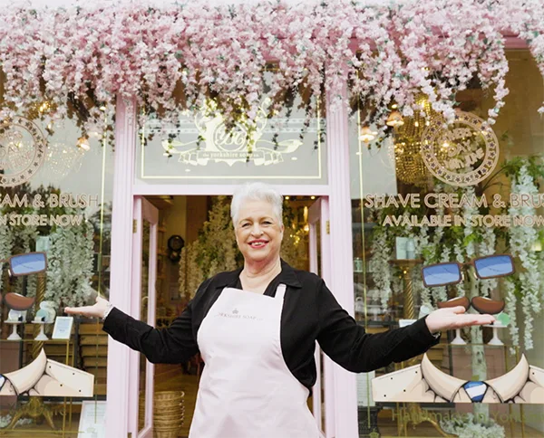 A middle-aged white woman with short silver/white hair wearing a pink apron over a black shirt, opens both arms out to the side in front of the open door of a pink-painted shop front. 