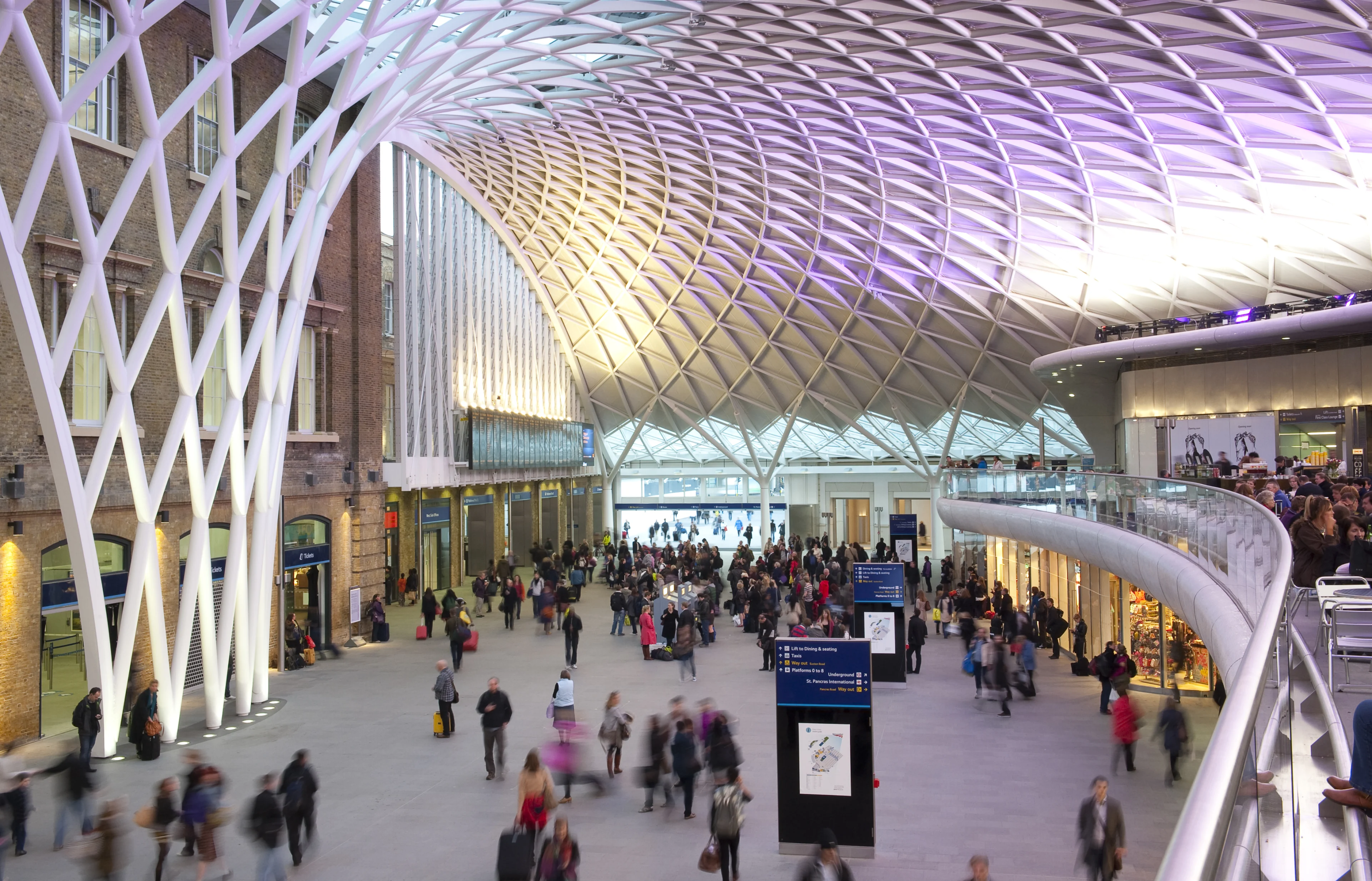 The concourse at Kings Cross station, showing passengers walking at the bottom level and seated on the balcony to the right hand side, with the large domed ceiling visible at the top. 