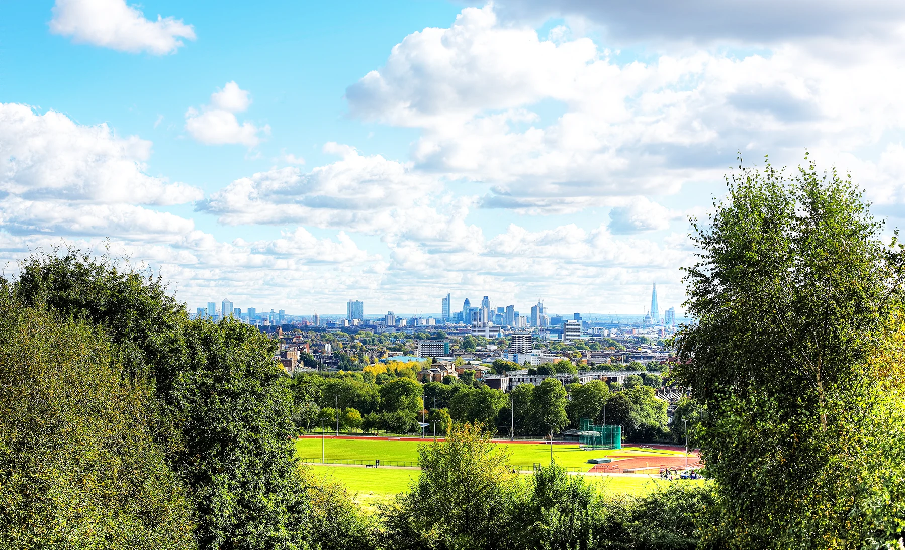 A panoramic view of London on a sunny day, looking over green fields to the skyline of tall towers in the background. 
