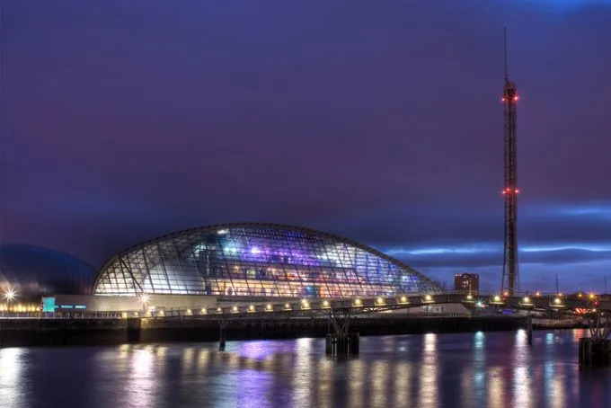 A arched riverside building made of metal and glass, lit up against a dark evening sky. 