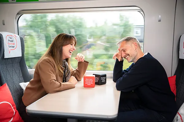 Actors Natalie Cassidy and Jack Ryder laughing together at a train table in "The Proper Catch-up Carriage", with conversation prompt dice between them.