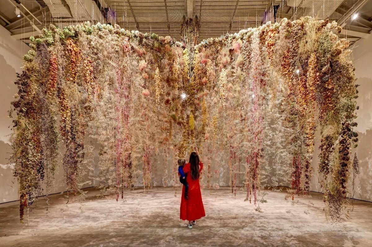 A woman holding a young child seen from behind and standing in front of a huge hanging installation of flowers and foliage at an art gallery.