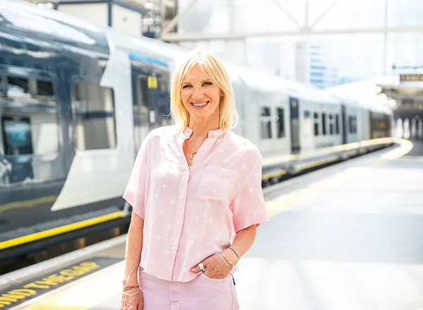 DJ Jo Whiley, a white woman with blonde hair, dressed in a summery pink blouse and trousers, smiling and standing on the platform of a railway station. 