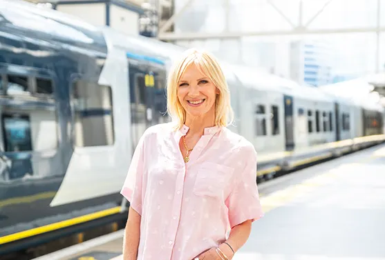 DJ Jo Whiley, a white woman with blonde hair, dressed in a summery pink blouse and trousers, smiling and standing on the platform of a railway station. 