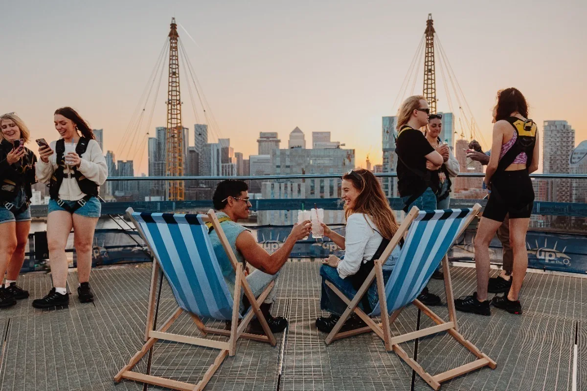 A man and a woman holding drinks and sitting in deck chairs on the roof of the O2 arena with the London skyline in the background and 2 small groups of people on either side. 