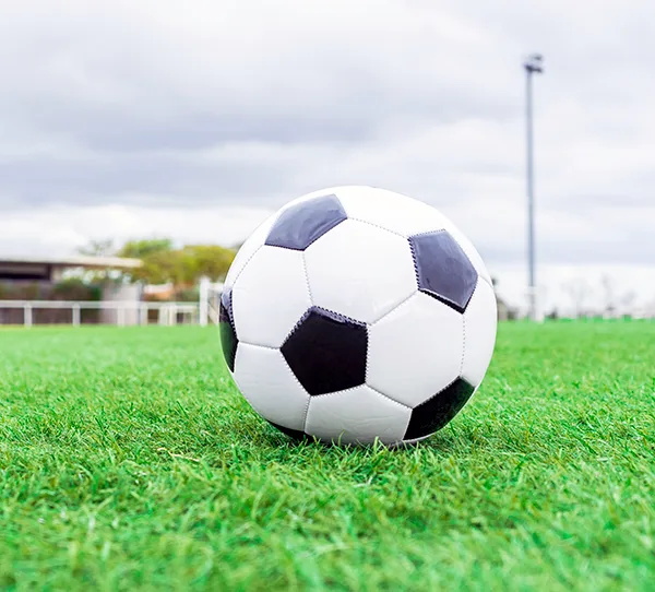 A black and white football ball on a green grass pitch with a goal, small stand and floodlights in the background. 