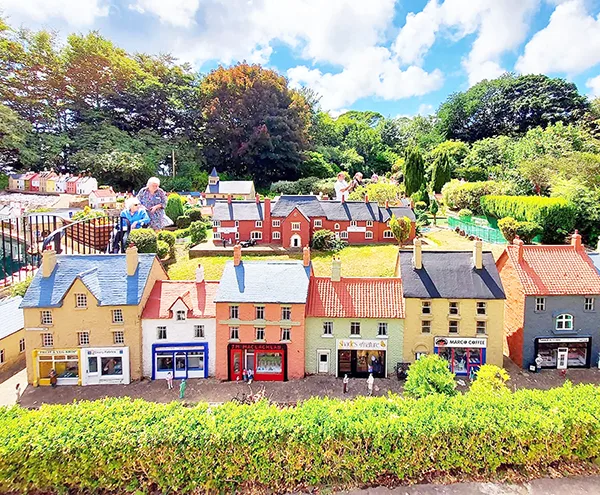 A detailed model village featuring colourful shops and houses, surrounded by lush greenery, with visitors viewing the display.