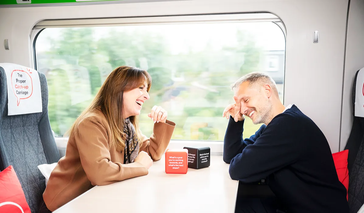 Actors Natalie Cassidy and Jack Ryder laughing together at a train table in "The Proper Catch-up Carriage", with conversation prompt dice between them.