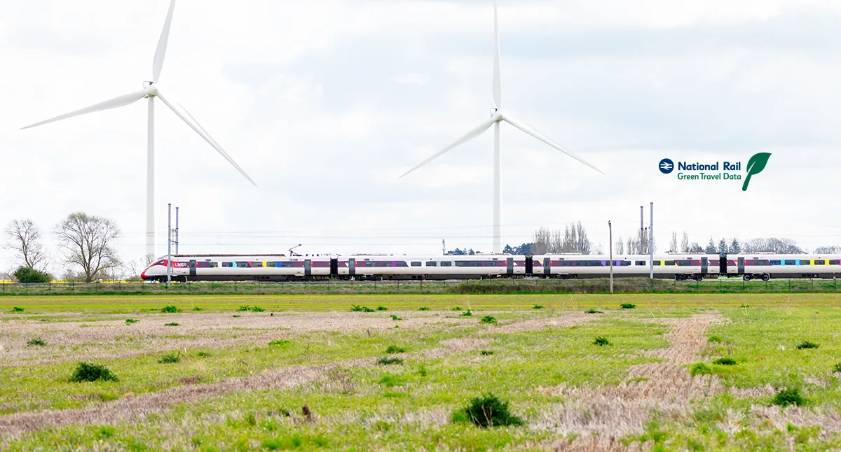 A train travelling across a field with large wind turbines in the background. 