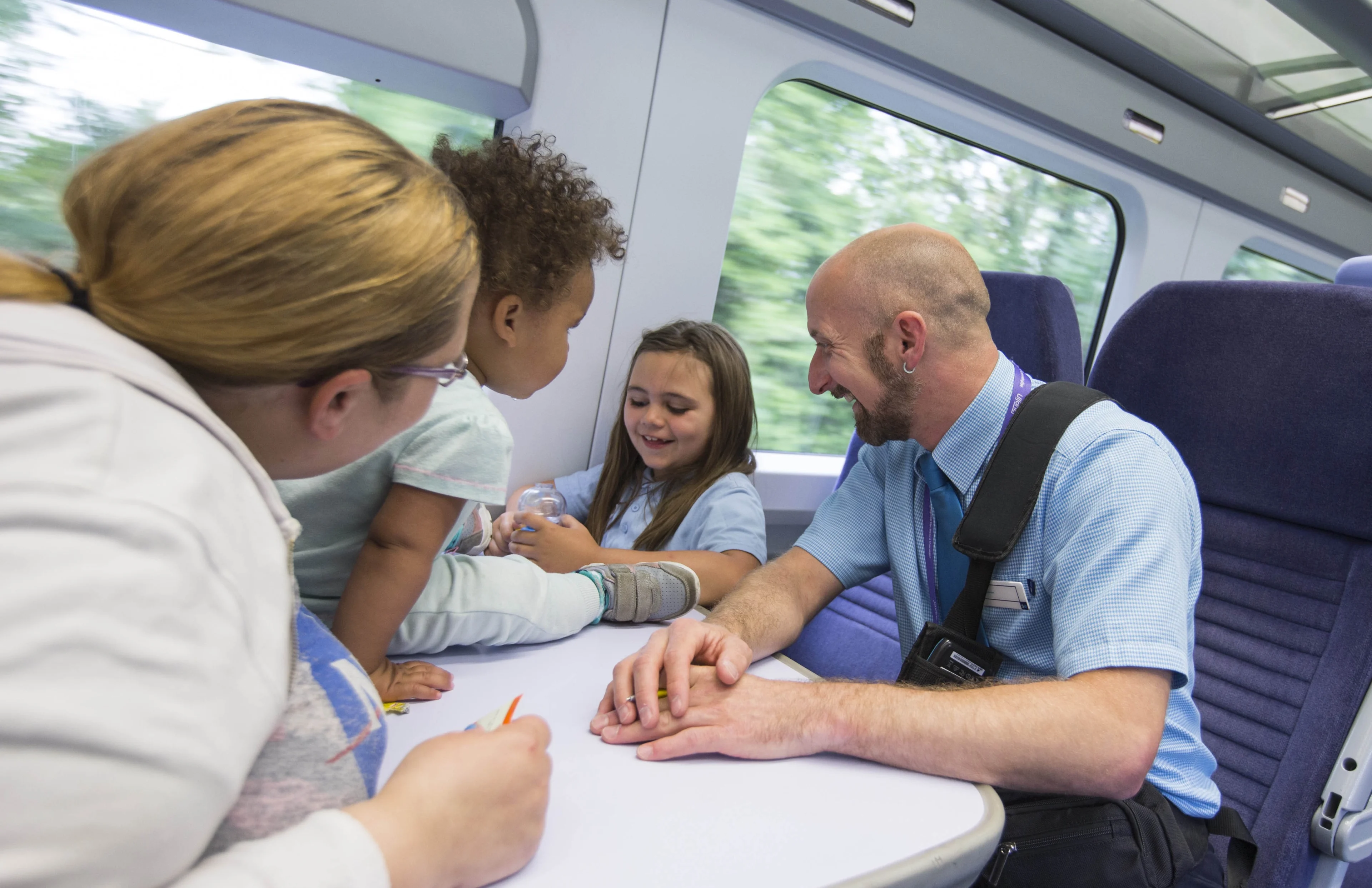 A woman and two children sit laughing at a table on a train with the onboard train guard, a man with a shaved head and beard.