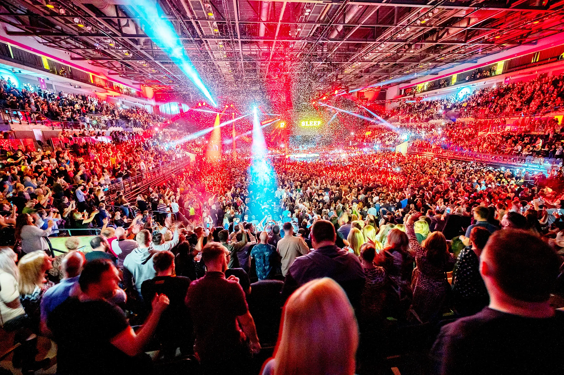 A crowd of people dancing at a concert in an arena with multicoloured lights.
