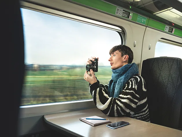 A white man with short dark hair sitting at a train table, he is holding a camera and looking out the window as the train goes past a green field.