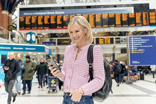 DJ Jo Whiley, a white woman with blonde hair, standing on the concourse of a busy railway station and looking at her phone.