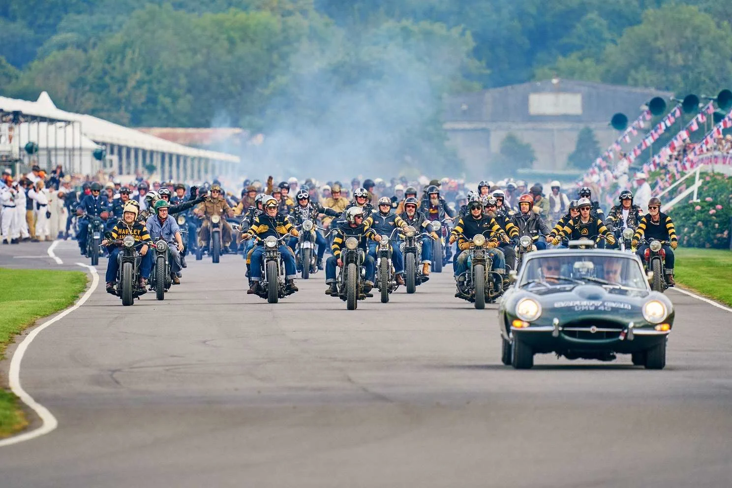 A vintage car being followed by lots of vintage motorcycles on a motor racing track. 