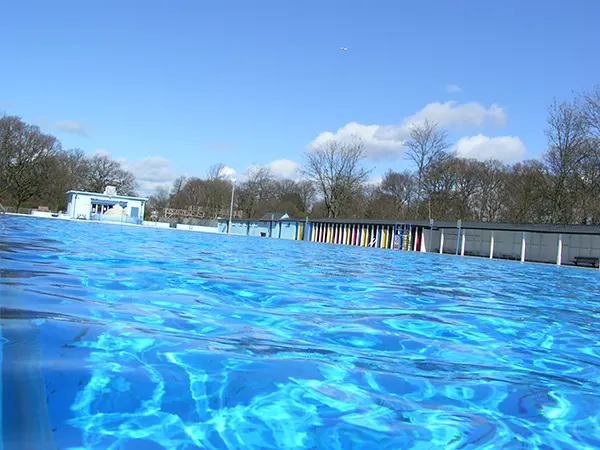 An outdoor lido with bright blue rippling water, colourful changing cubicles, bare trees, and a clear blue sky.