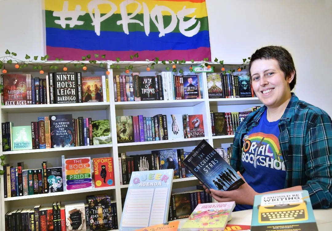 A smiling white person with short hair holding a book and standing in front of displays in a bookshop, with a rainbow banner that says PRIDE on the wall behind. 