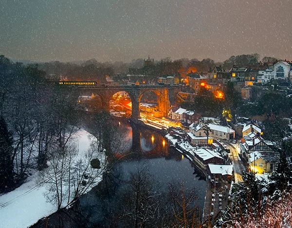 Snowy night view of a train crossing an illuminated viaduct over a river, with houses with warm lights in winter on the banks. 