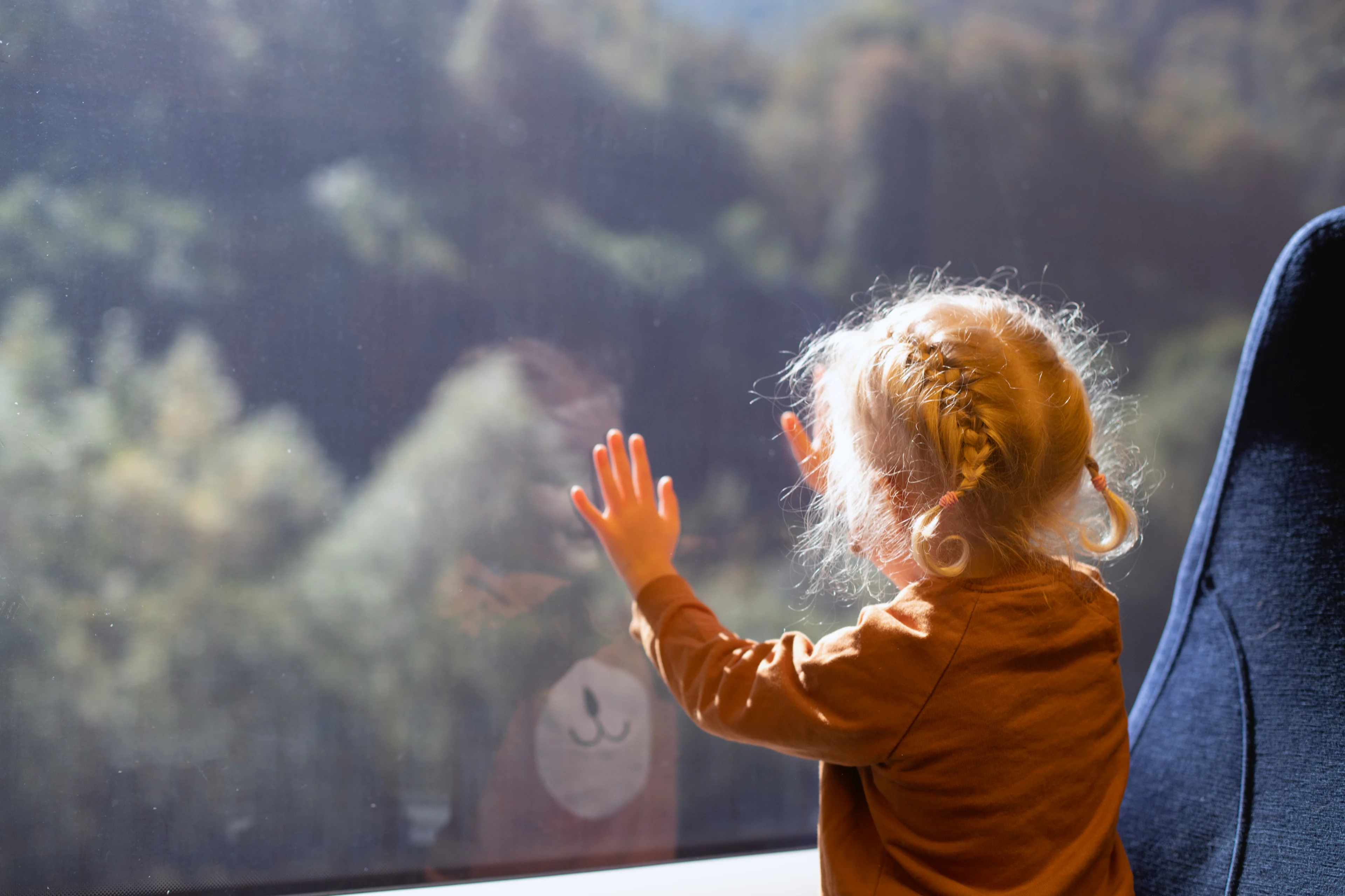 A young girl is reflected in a train window as she puts her hands on the glass to look out at the landscape