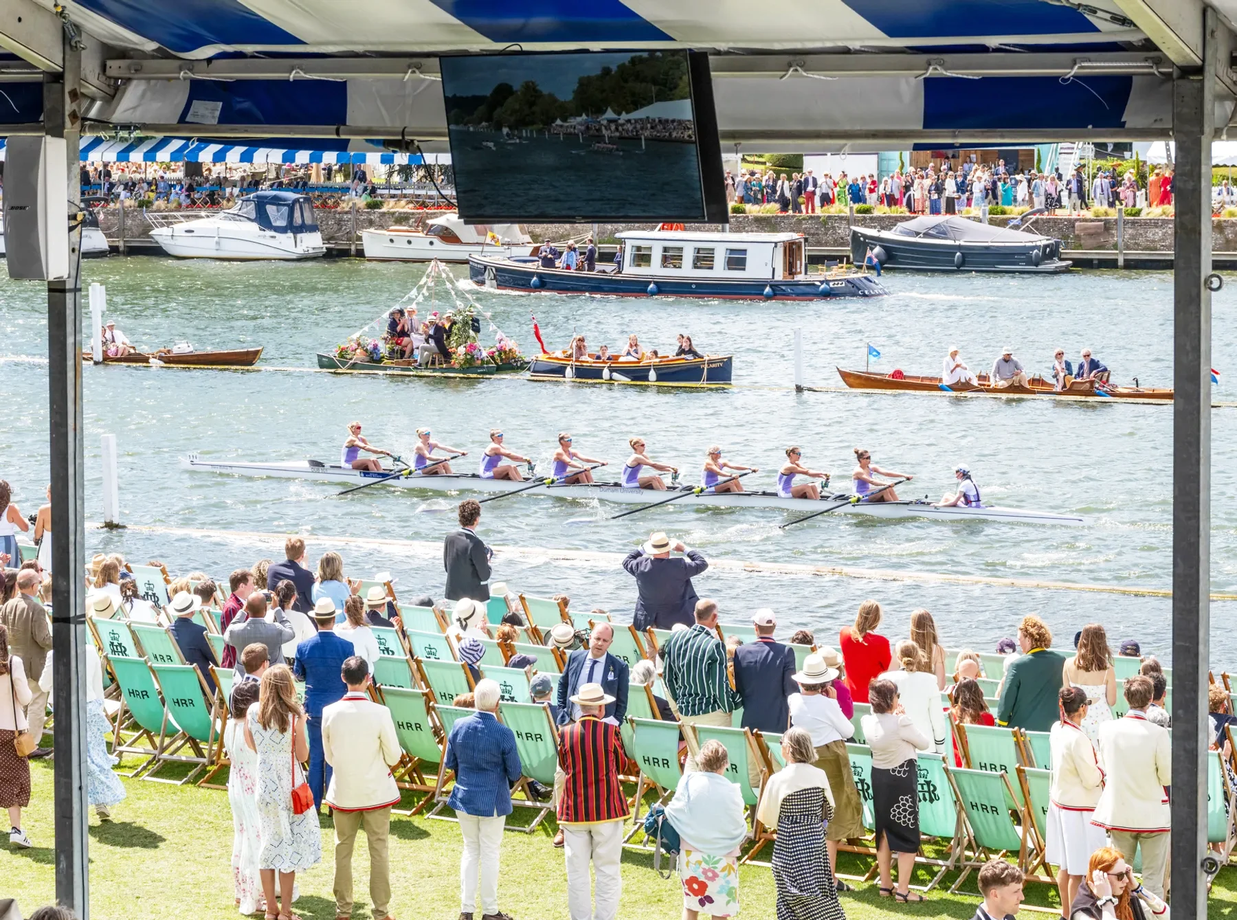 Teams rowing on a river with people standing and in deck chairs on the banks watching them. 