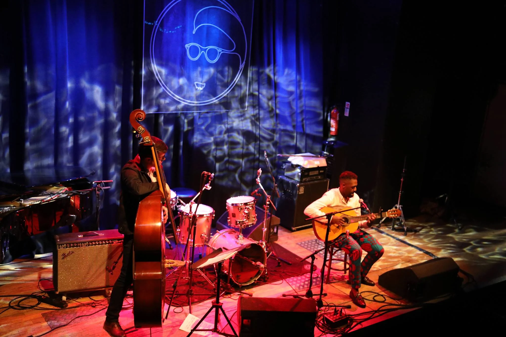 A group of Black musicians playing guitar, double bass and drums on the stage of a small venue. 
