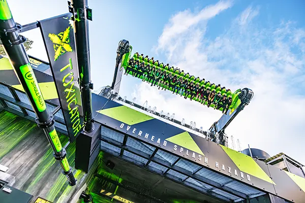 A theme park ride called the Toxicator with a long, horizontal row of green seats carrying passengers, suspended by two large mechanical arms.