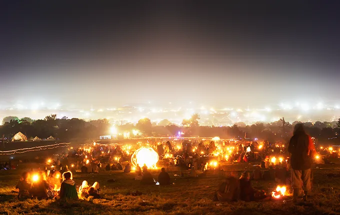A field at dusk full of festival-goers with camp fires and the lights of Glastonbury in the distance.