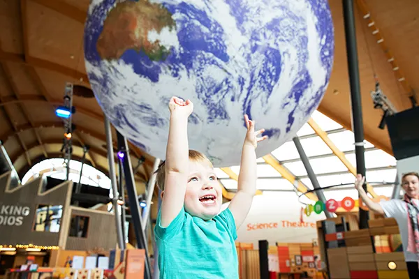 A white toddler holding their arms up in the air in front of a huge hanging globe of the Earth. 