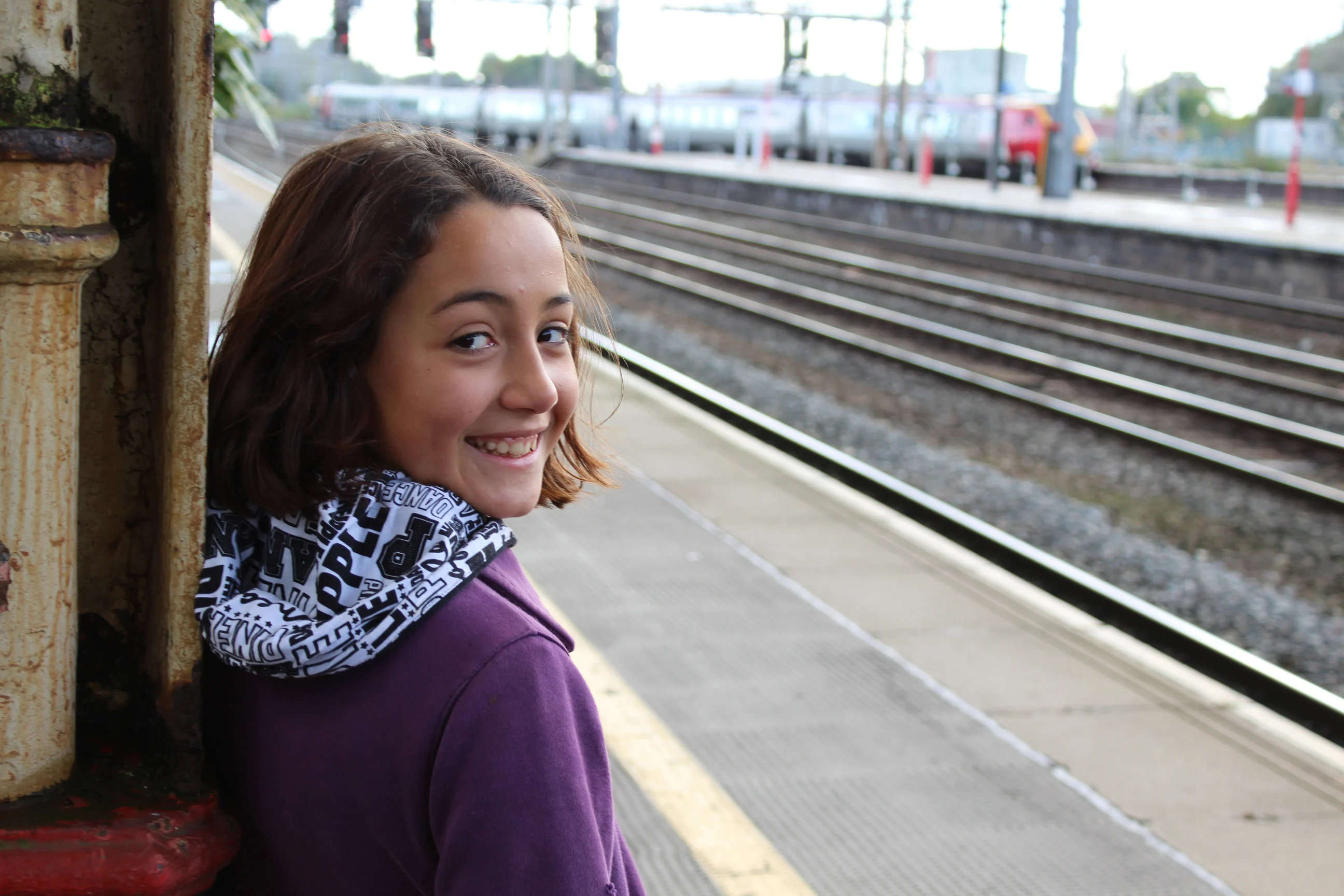 A girl in a purple top and with a black and white hood stands on a railway platform, smiling at the camera. 