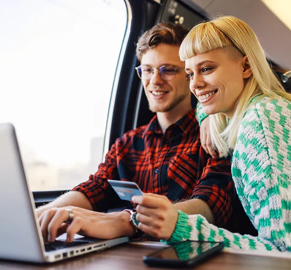 A young white man with a check shirt and glasses, and a white woman with blonde hair and a blunt fringe, sitting at a train table and using a laptop and credit card to do online shopping. 