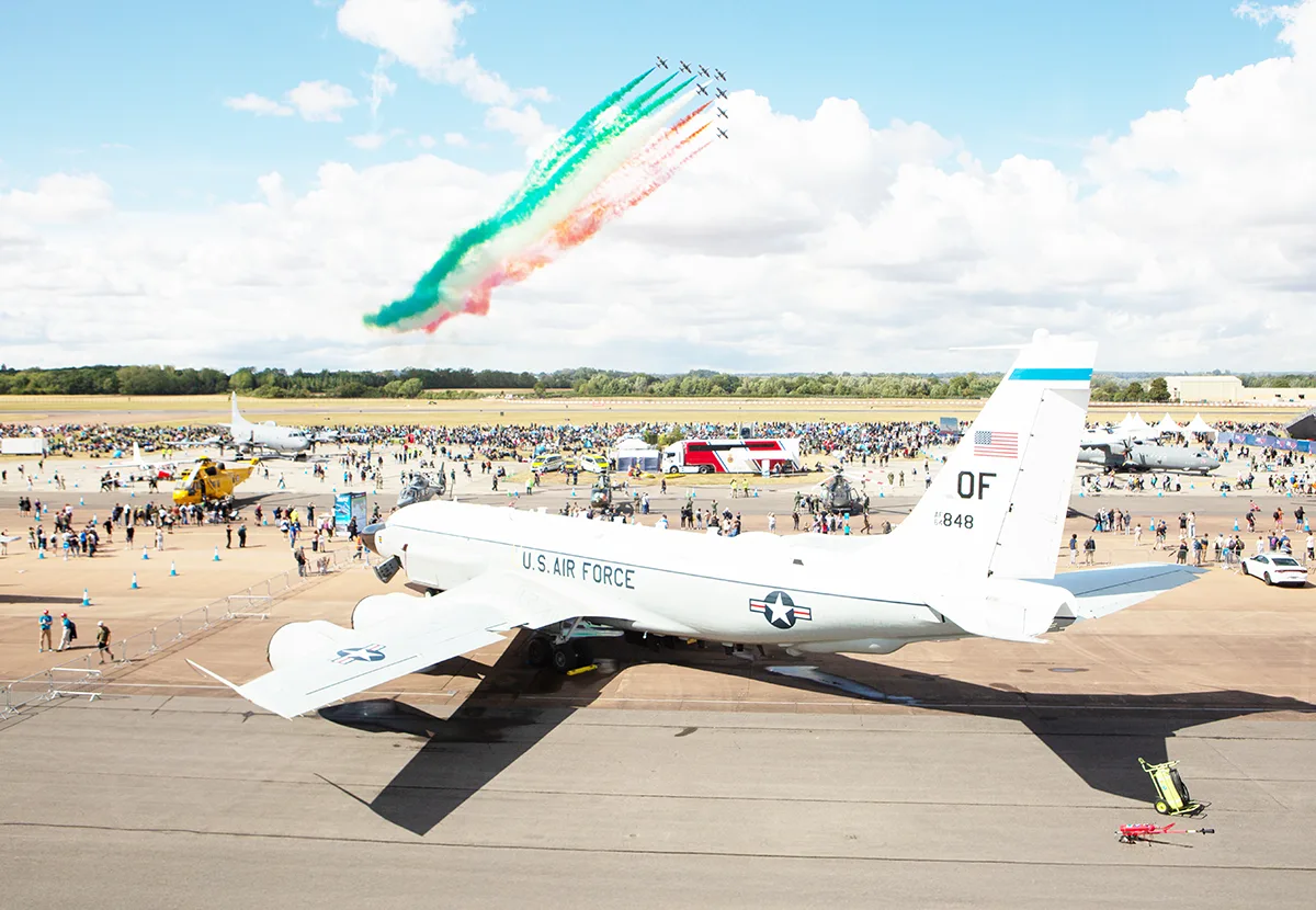 A U.S. Air Force aircraft on the tarmac at an airshow, with display jets trailing green, white and red smoke overhead.