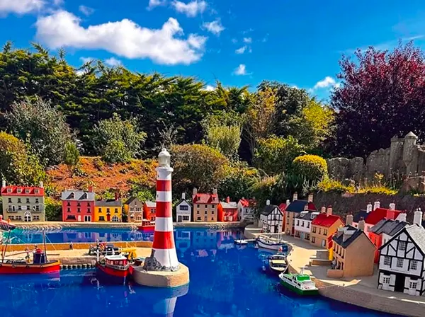 A model village featuring a harbour, striped lighthouse, colourful buildings, and small boats on blue water, surrounded by trees.