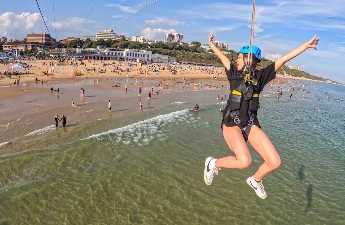 A young white woman wearing a helmet and harness dangles over the sea at an adventure resort.