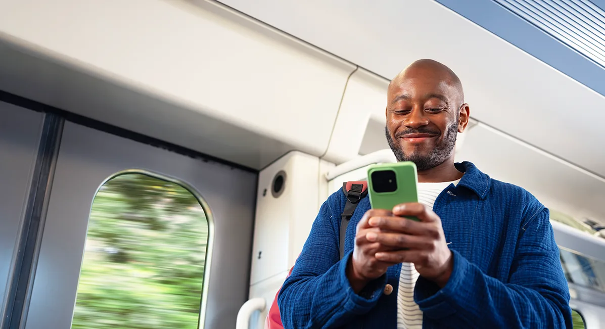 A smiling black man with a bald head and beard, standing on a train and looking at his mobile phone. 