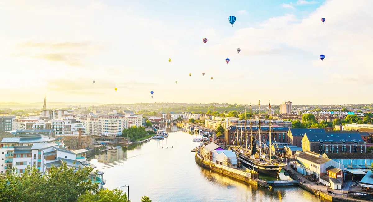 Multicoloured hot air balloons floating above a river running through a city.