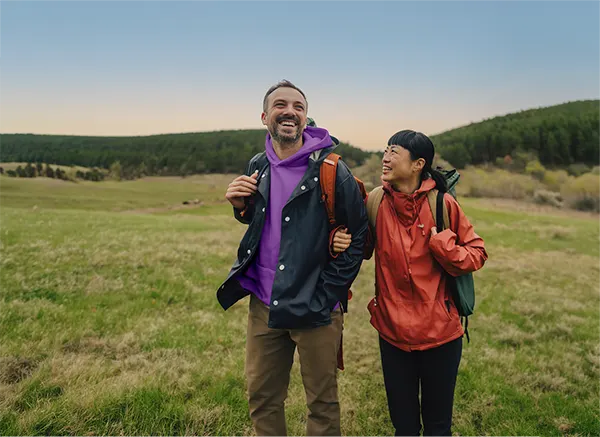 Two smiling hikers with backpacks stand in a green meadow, with a forest and clear sky in the background.