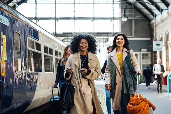 A group of smiling female friends on a station platform, a Black woman with long natural hair and a South Asian woman with long hair are at the front carrying luggage.