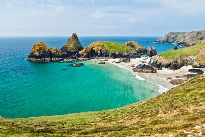 Kynance cove sits with white and grey sand with green island esque pieces of land sitting on a sea of green and blue hues beneath a hazy blue sky. Green grass sits in foreground.