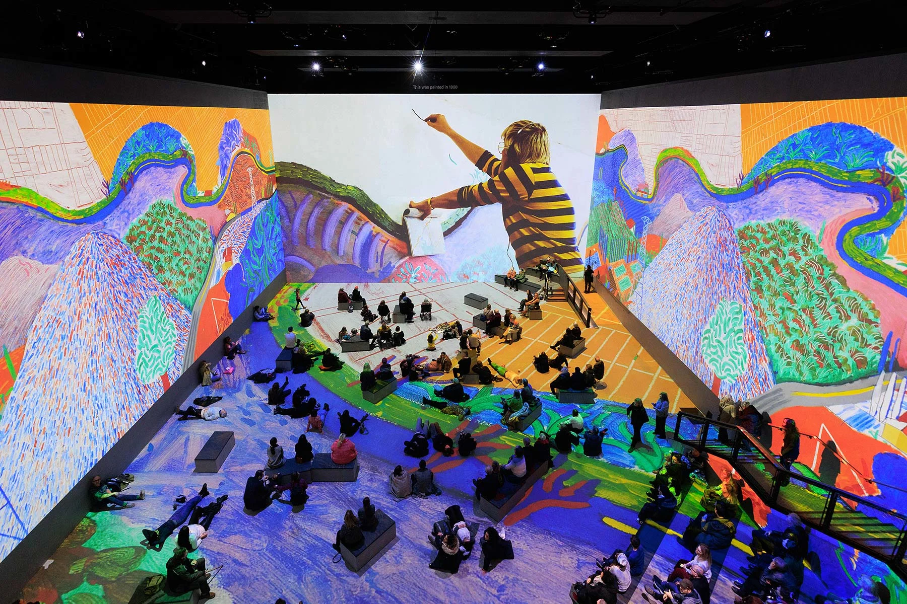 People sitting in a cavernous room with projections of paintings on the walls and floor, and a large screen showing the artist David Hockney at work
