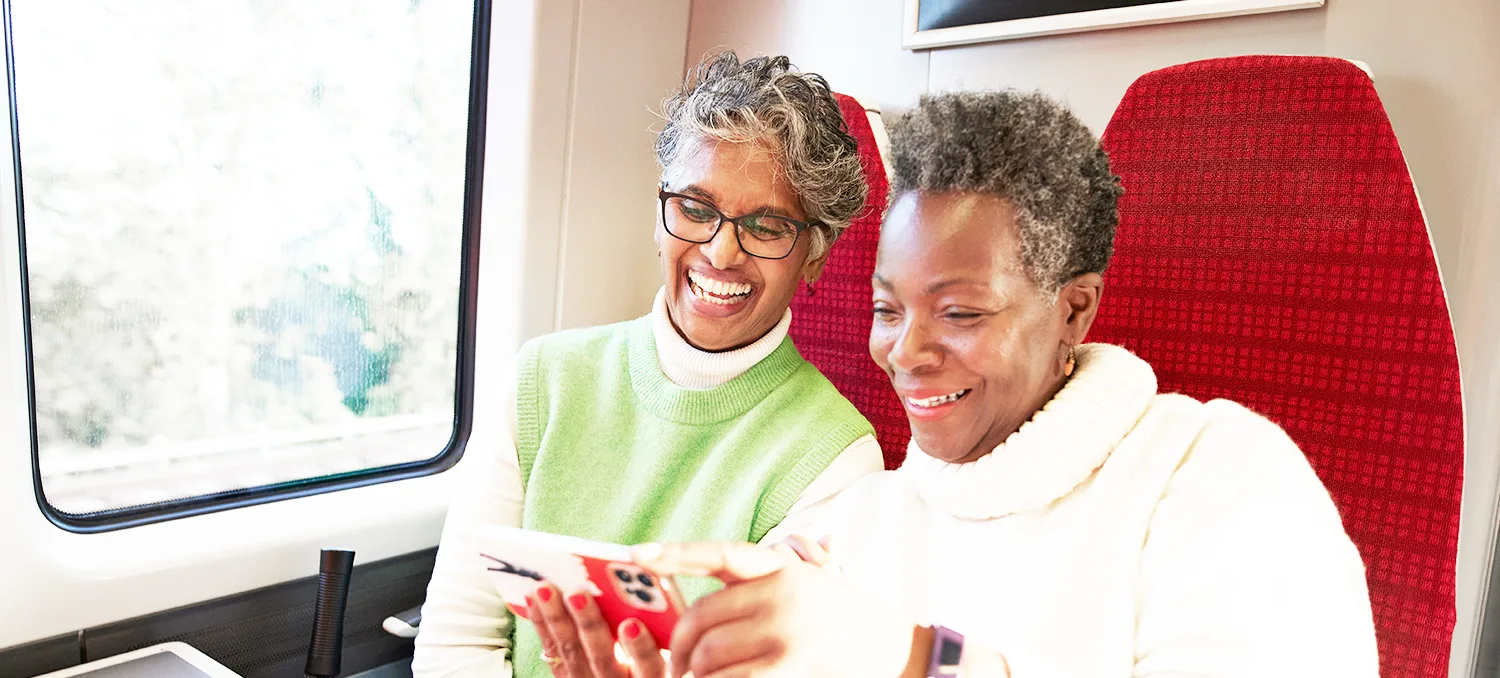 2 senior women laughing and smiling sitting at a train table looking at a smartphone