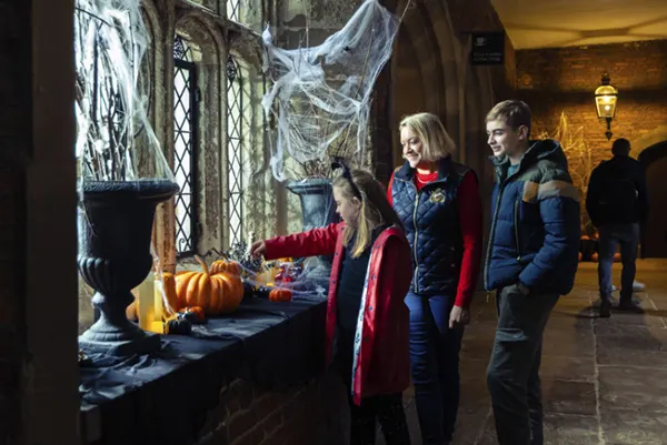 A family exploring Halloween decorations including pumpkins and cobwebs by a leaded window in a dimly lit historic building.