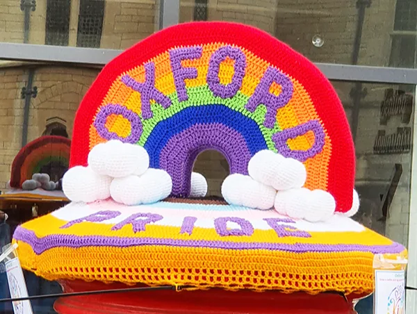 A red post box with a knitted cover on top that has a rainbow and says OXFORD PRIDE in purple letters.