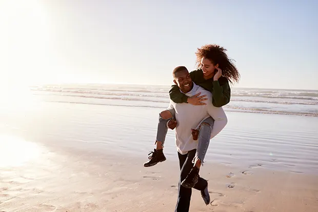 A Black man gives a Black woman a piggyback as they walk along a sandy beach. 