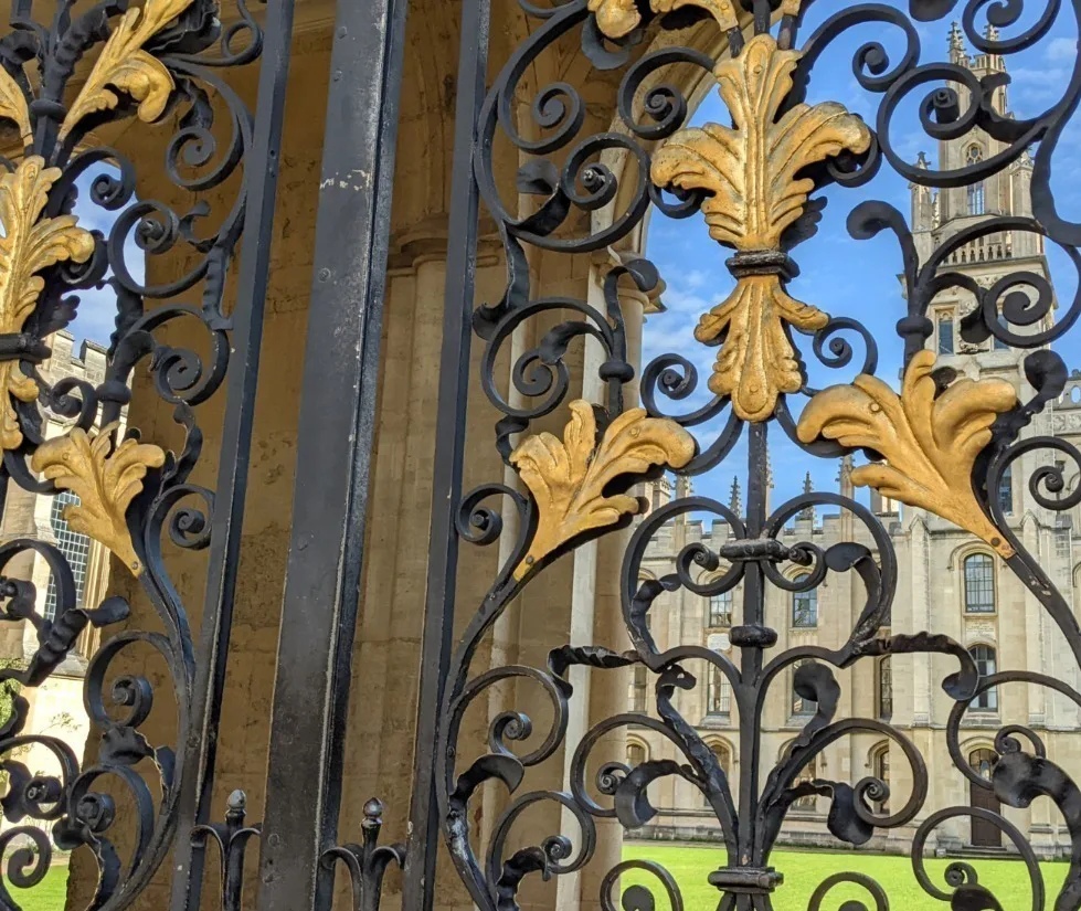 Looking through wrought iron and gilded metal gates onto an Oxford university building and lawn. 