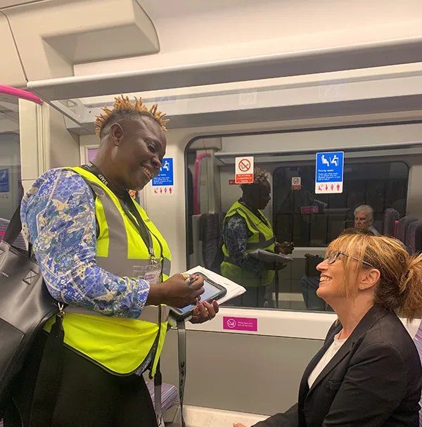 Black woman in high-vis vest conducting a survey on a train carriage, using a tablet, while smiling at a female passenger.