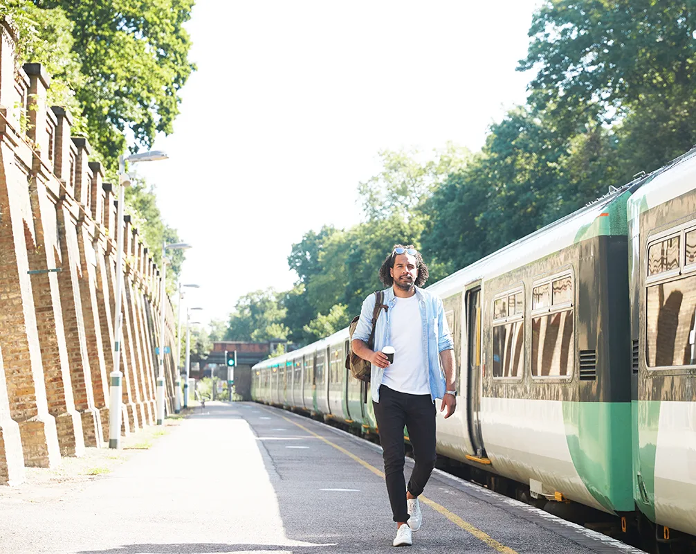 Person walking along sunny train platform with coffee cup, wearing backpack beside green and white train.