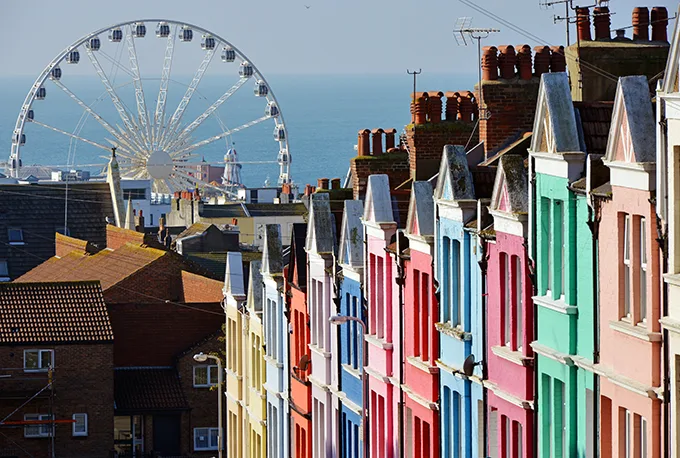 A row of brightly painted terraced houses with a view of the sea and a ferris wheel in the background.